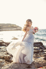 Fototapeta na wymiar Beautiful young bride in long white wedding dress,beautiful long curly blonde hair,bride posing alone,standing on the rocky shore against the clear blue sky and ocean in summer time in fresh air