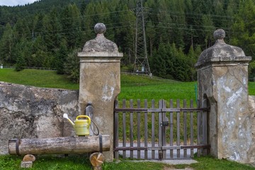 Holzbrunnen mit Gießkanne an Friedhofsmauer