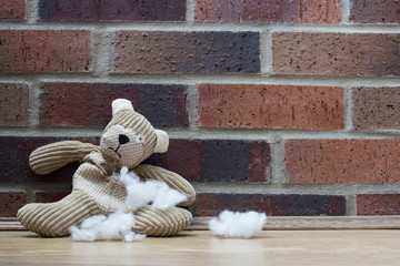 A sad, abandoned teddy bear with stuffing and filling falling from a hole in his ripped and torn tummy and sitting alone against a brick wall. - obrazy, fototapety, plakaty