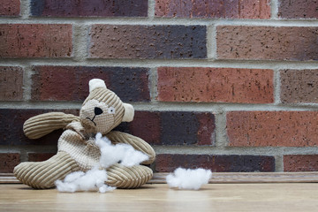 A sad, abandoned teddy bear with stuffing and filling falling from a hole in his ripped and torn tummy and sitting alone against a brick wall.