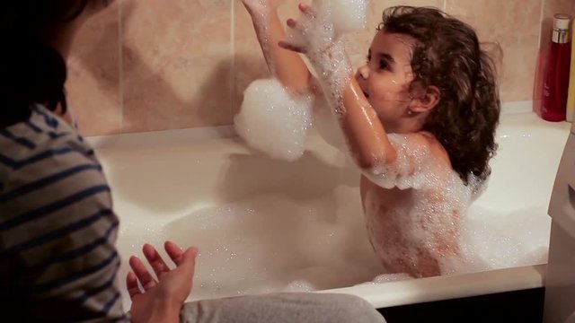 Beautiful curly-haired girl bathing in the bath with foam. Playing with foam bath. Mother bathes charming daughter