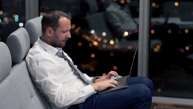 Angry businessman destroying laptop sitting on sofa at home at night
