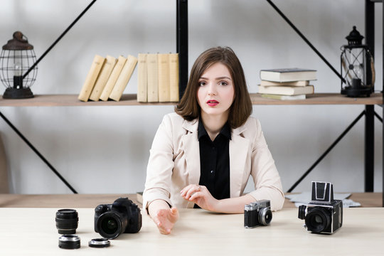 Blogger reasoning about photo equipment. Young beautiful brunette talking about photographer work with different cameras on table