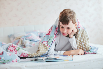 Fototapeta na wymiar boy reads a book lying under a blanket on the bed
