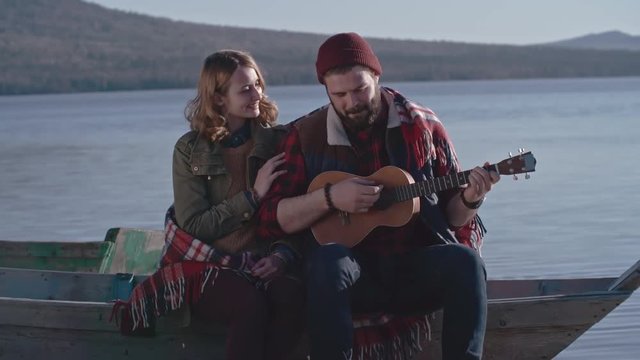 Lockdown with slow motion of young bearded man in beanie hat sitting on moored boat near lake and playing mini acoustic guitar to his affectionate girlfriend 