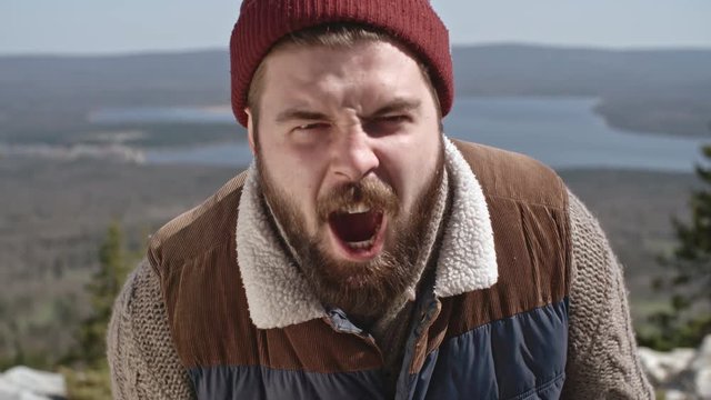 Slow motion medium shot of young man with beard sitting on mountain ledge screaming in rage and looking at camera