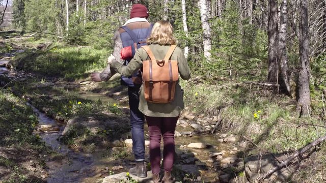 Handheld shot of bearded boyfriend jumping over brook in forest and helping his blond girlfriend during hiking trip