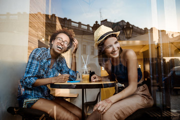 Two young beautiful girls smiling, laughing, resting in cafe. Shot from outside.
