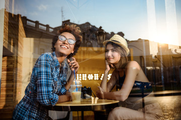Two young beautiful girls smiling, speaking, resting in cafe. Shot from outside.
