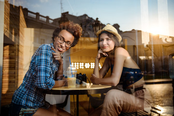 Two young beautiful girls smiling, speaking, resting in cafe. Shot from outside.