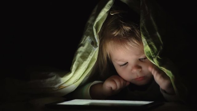 Concept of technology and lifestyle in baby life. Little girl using tablet pc under blanket at night. 4K video