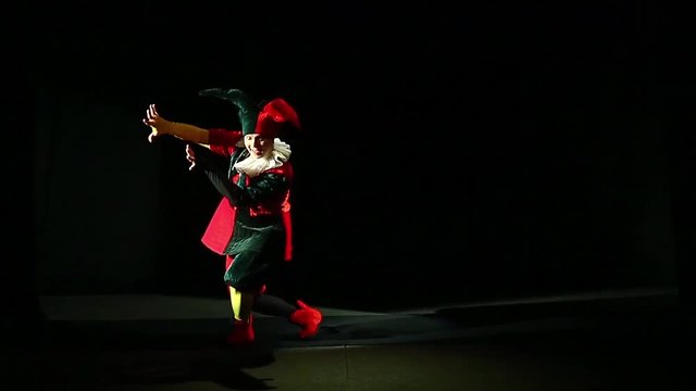 a man dressed in a jester costume is playing on a black background and exits. overall plan
