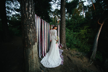 Tanned brunette in a long wedding dress stands in the dark fores
