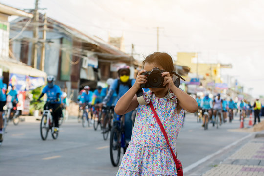 Adorable little girl taking a photo by camera with cyclists ride on road background.