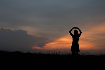 silhouette of woman heart shape making of hands against at sunse