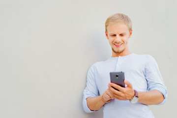 Youth and technology. Handsome young bearded man using smart phone and smiling while standing against grey wall.