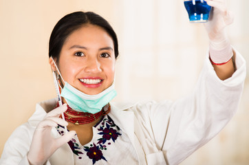 Young beautiful woman dressed in doctors coat and red necklace, facial mask pulled down to chin, holding up blue laboratory accessory, smiling happily, egg white clinic background