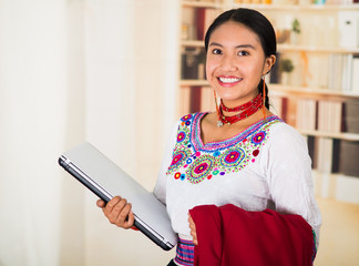 Beautiful young lawyer wearing traditional andean blouse with necklace, holding laptop and red jacket smiling, bookshelves background