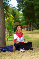 Young woman wearing traditional glasses, andean skirt and blouse with matching red necklace, sitting on grass next to tree in park area, relaxing while reading book, smiling happily