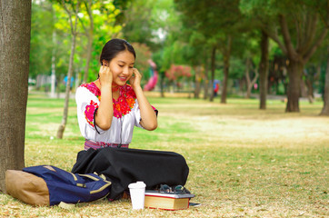 Young woman wearing traditional andean skirt and blouse with matching red necklace, sitting on grass next to tree in park area, relaxing while using mobile phone headphones connected, smiling happily