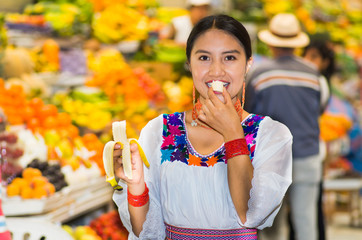 Beautiful young hispanic woman wearing andean traditional blouse posing for camera while eating banana inside fruit market, colorful healthy food selection in background