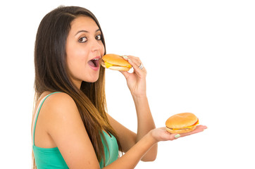 Beautiful young woman posing for camera eating hamburger while holding another one in other hand, smiling happily, white studio background
