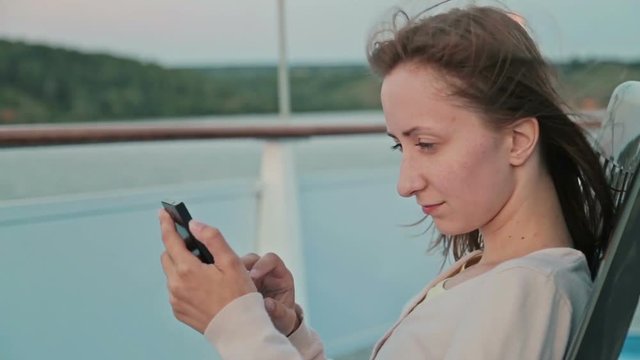 Young beautiful woman standing on deck of cruise ship and using her smart phone. After sunset. Strong wind waving her hair. The river and riverside can be seen in the background behind her