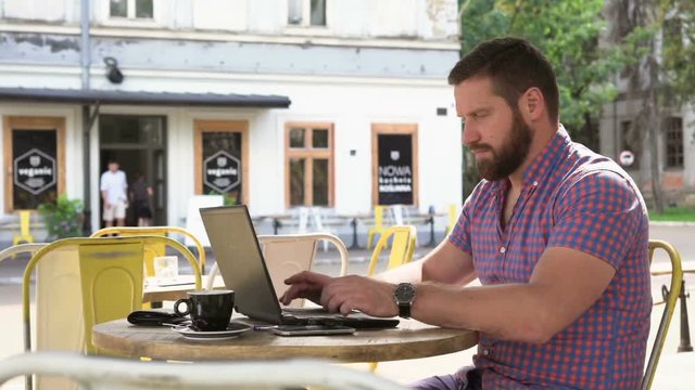 Young man writing on laptop. He is sitting at the table in cafe. He is young and has beard. Man is dressed in checkered shirt. Slider shot right
