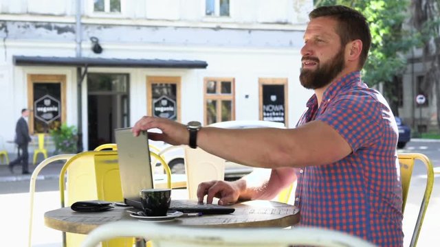 Young man ends typing on laptop and relaxes. He is sitting at the table in cafe. He is young and has beard. Man is dressed in checkered shirt. Slider shot right
