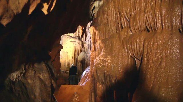 We see moisten wall of stalactites in a cave, that expand all down to the path a tourist is walking on. There is a narrow pass on from the wall. Wide-angle shot.

