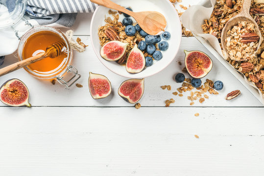 Healthy breakfast set. Oat granola with nuts, yogurt, honey, fresh figs and blueberries in bowl on white wooden background, top view, copy space