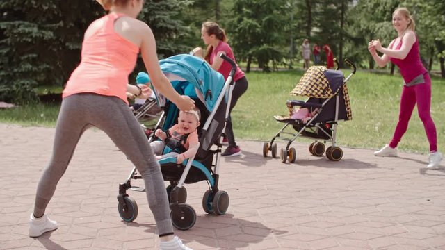 Group of women standing in front of their babies in strollers and doing side lunges with fitness instructor showing exercise and playing with her adorable baby