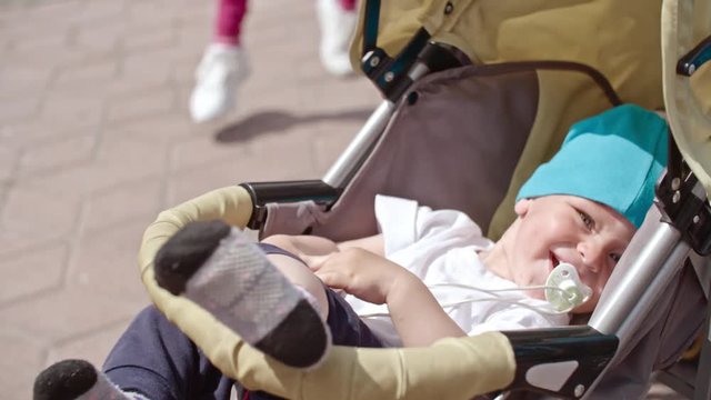 Cute little boy sitting in stroller with pacifier and laughing while his mother tickling him during squatting 