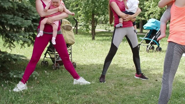 Two smiling young women doing squats with cute little babies on their arms with fitness instructor on green lawn in the park