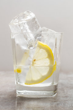 Vaso con agua y una rodaja de limón y hielo sobre fondo gris. Vista de frente