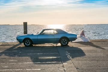 Fototapeta na wymiar Eine Braut sitzt alleine neben dem Auto und beobachtet Sonnenuntergang am Meer