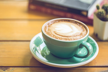 hot latte art in green cup on wooden table with book and tree.