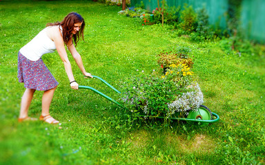 young woman in garden playfully pushing decorative wheelbarrow full of flowers.