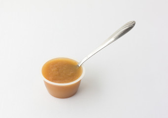 Cinnamon applesauce in a plastic container with a spoon atop a white tablecloth.