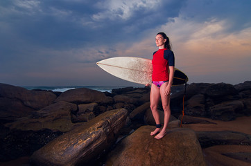 Young surfer woman girl with surf board standing on rocks looking at ocean 
