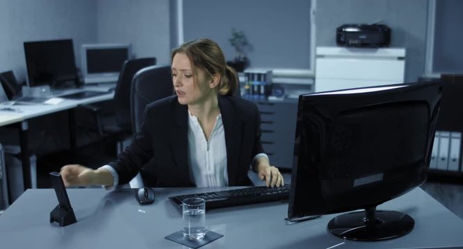 4K: Eine junge Angestellte sitzt angespannt an ihrem Arbeitsplatz. Ein Telefonat st&ouml;rt ihre Computerarbeit. Sie trinkt einen Schluck Wasser und ist sehr ver&auml;rgert.