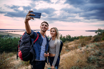 Young caucasian couple making selfie while hiking outdoors with backpacks during sunset 