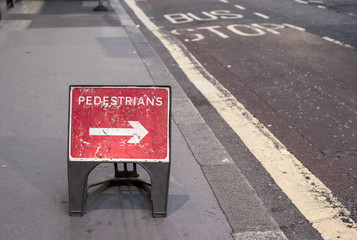 a red 'pedestrians' sign in the middle of the sidewalk, with an arrow pointing towards the road