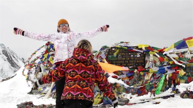 Two very happy women congratulate each other, hug and jump on the top of high snowy mountain after difficult climbing expedition in front of buddhist praying flags