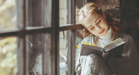 Happy young woman reading book by window in fall