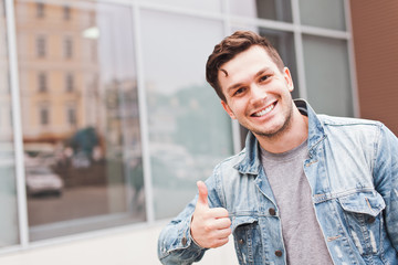 Young successful handsome smiling man businessman in casual jeans jacket showing thumbs up gesture outdoors on street in summer in front of mirror glass office building or business center