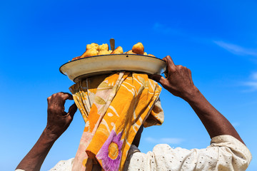 Woman carrying a dish with food on her head seen from behind - obrazy, fototapety, plakaty