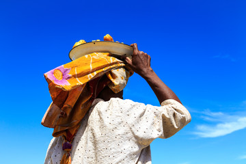 Woman carrying a dish with food on her head seen from behind - obrazy, fototapety, plakaty