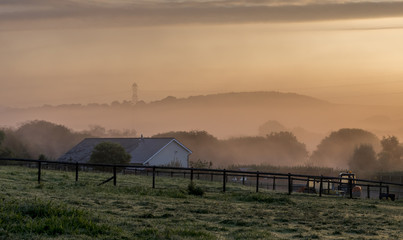 Autumn dawn, with an orange light through the early morning mist, looking over a rural farm scene to distance hills