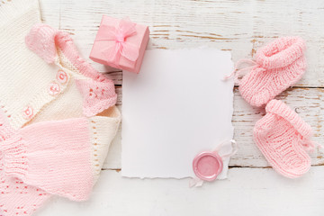 Baby girl clothes, blank card withwax seal and small present box on white wooden background. Flat lay. Owerhad view